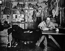 Man sitting cross-legged at table, pipe in hand, apparently writing. Much clutter of clothing, books and equipment is in the background.