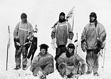 Five men(three standing, two sitting on the icy ground) in heavy polar clothing. All look exhausted and unhappy. The standing men are carrying flagstaffs and a Union flag flies from a mast in the background. Scott's party at the South Pole.  Left to right: Oates; Bowers; Scott; Wilson; Evans