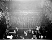 A photo taken from a ceiling of a tall square industrial room. Cement walls have metal ladders and meshes. A dozen of people are engaged in some activities on the floor.